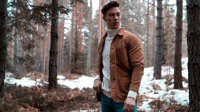 男人穿着白色高领毛衣和棕色夹克站在森林
